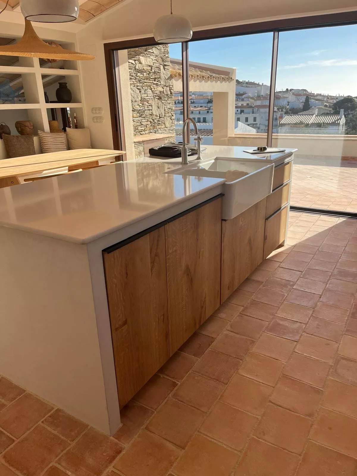 Cocina moderna con isla central, encimera blanca, muebles de madera clara y fregadero de cerámica, vistas al mar y pueblo.