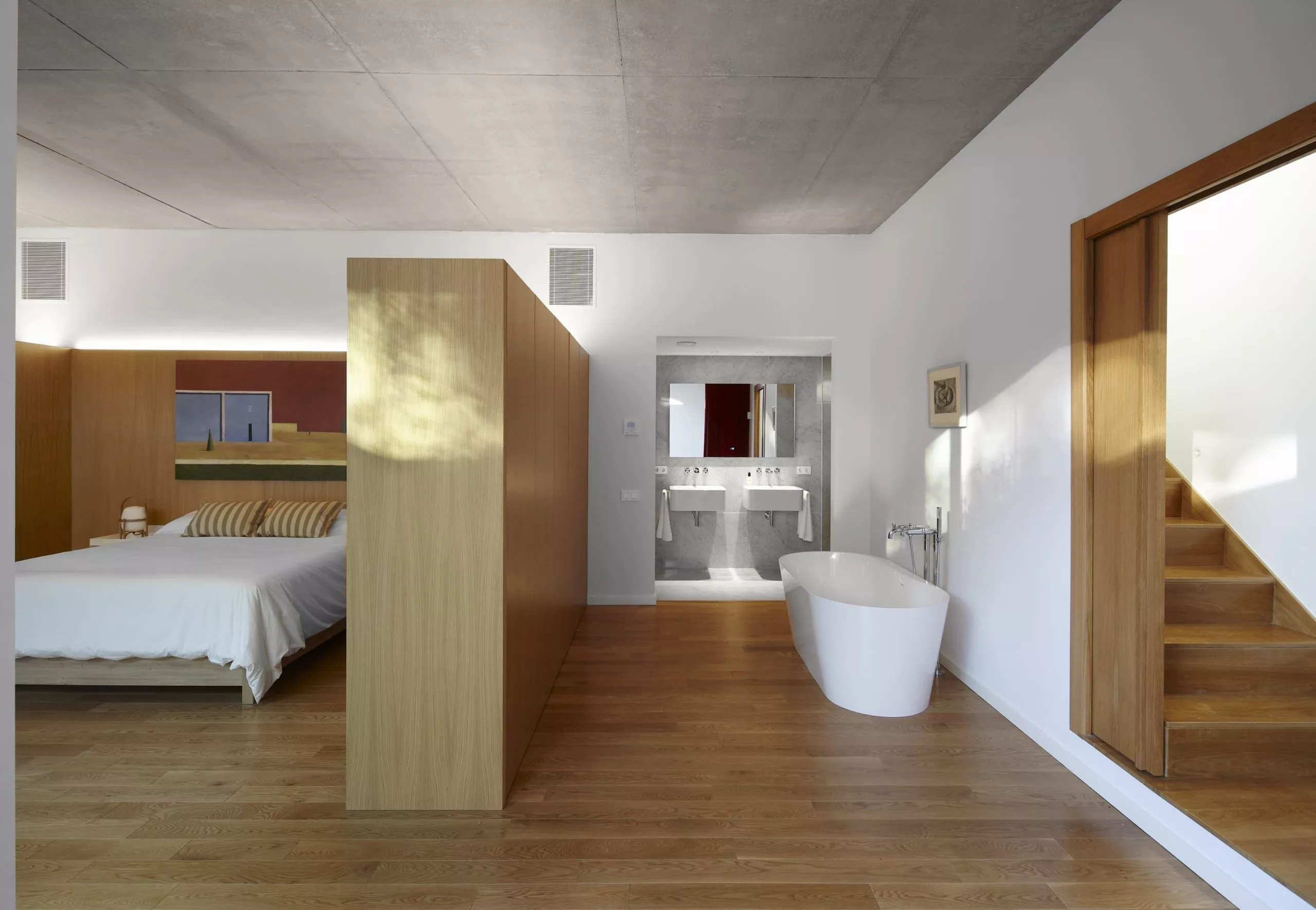 Dormitorio moderno con cama blanca, cabecero de madera, baño integrado con bañera y lavabos dobles, y escaleras de madera.
