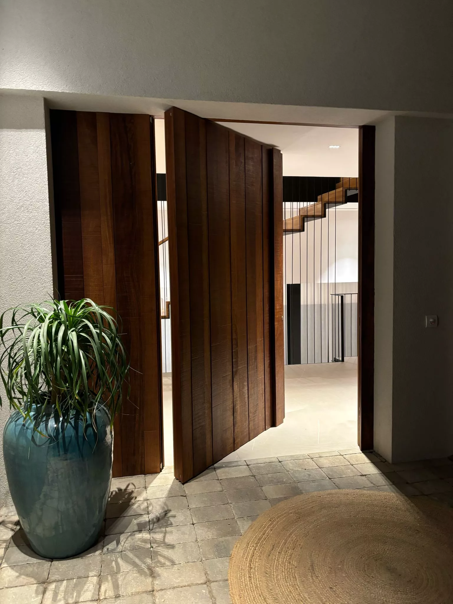 Entrada moderna con puertas de madera maciza, maceta azul verdoso y alfombra de yute, mostrando carpintería a medida.