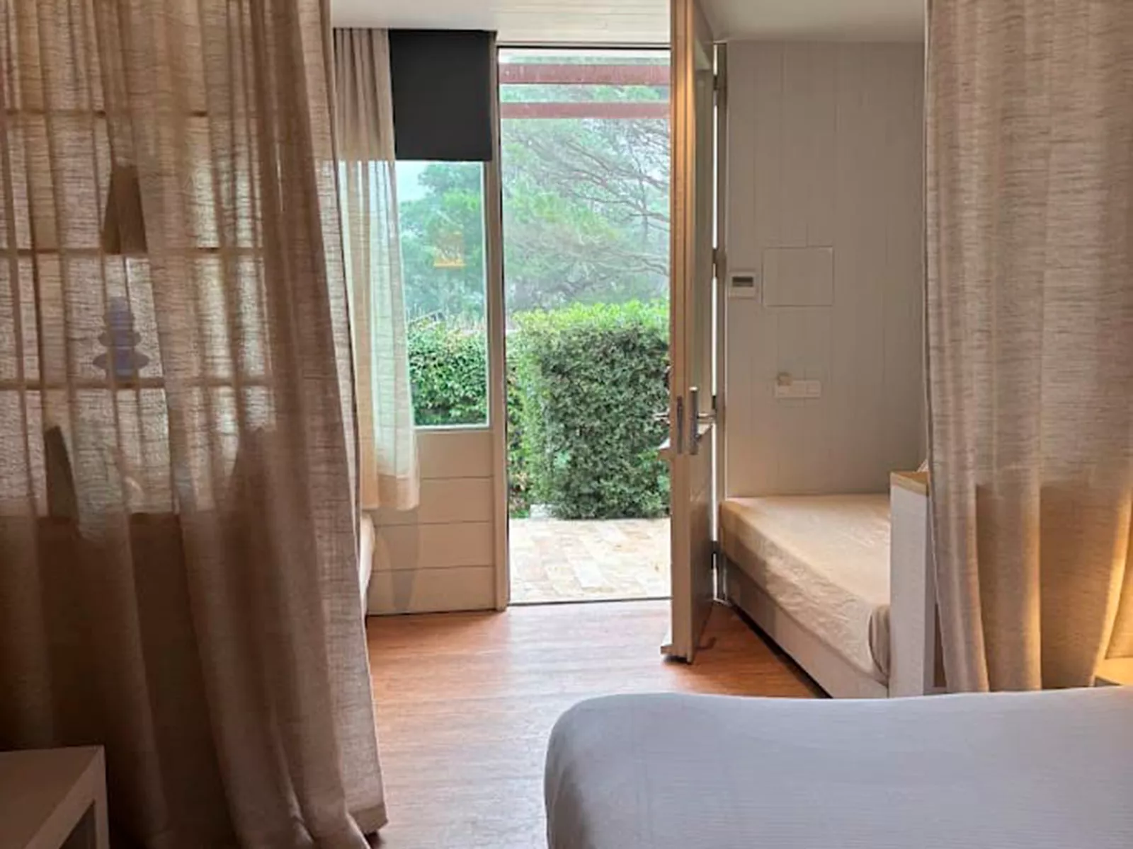 Habitación de hotel con cama individual, cortinas de lino y puerta abierta a un jardín verde, mostrando carpintería a medida.