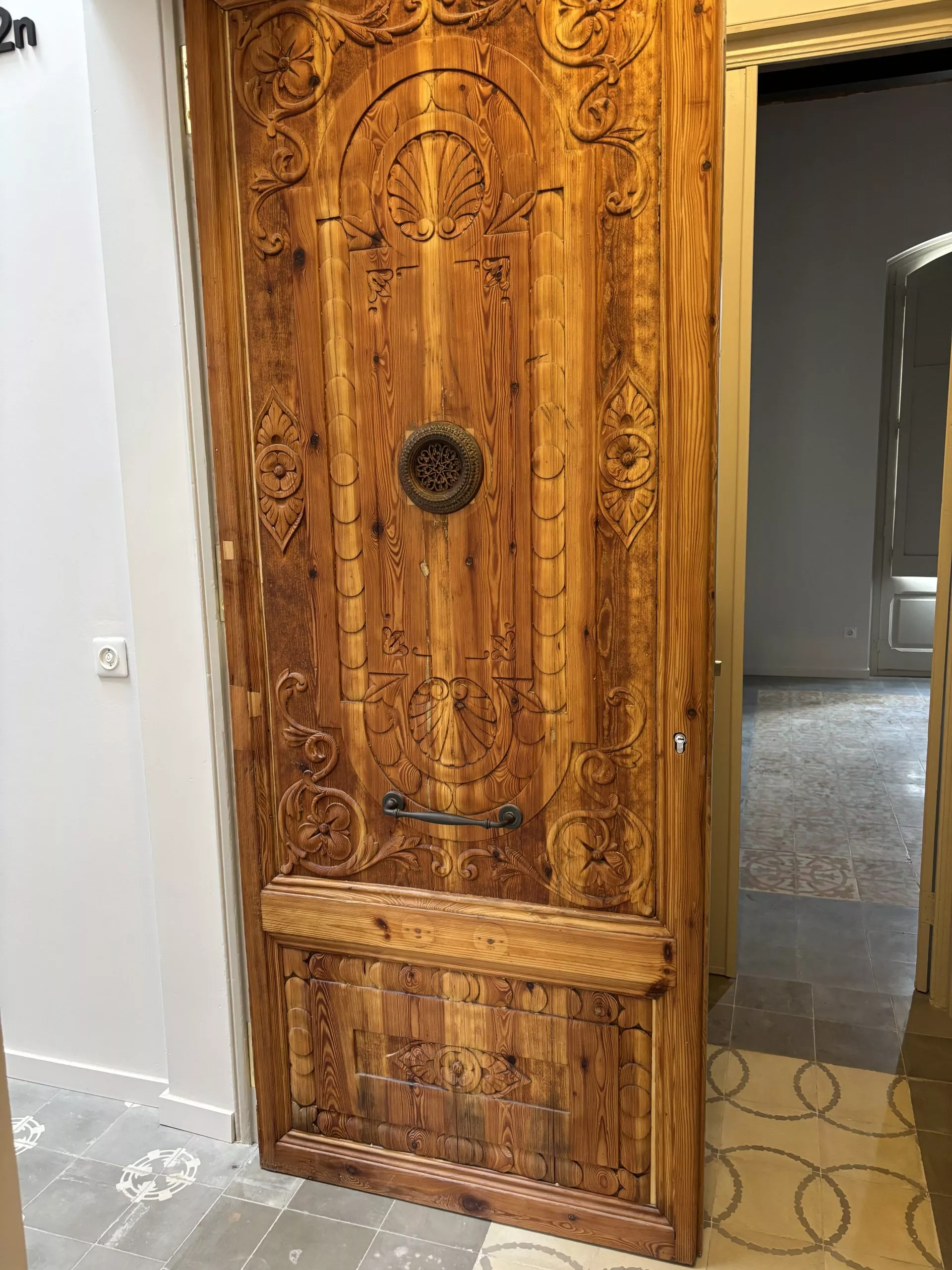 Una porta de fusta tallada a mà amb detalls ornamentals i ferratges antics, en una casa amb sòls de rajoles.