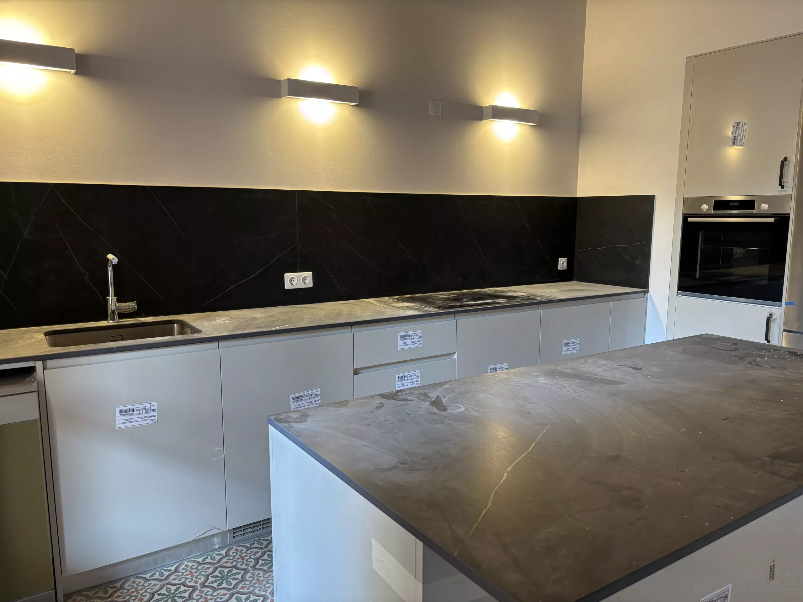 Cuina moderna amb taulell de marbre fosc, rentamà, aixeta cromada, placa d'inducció i forn integrat, il·luminació càlida.
